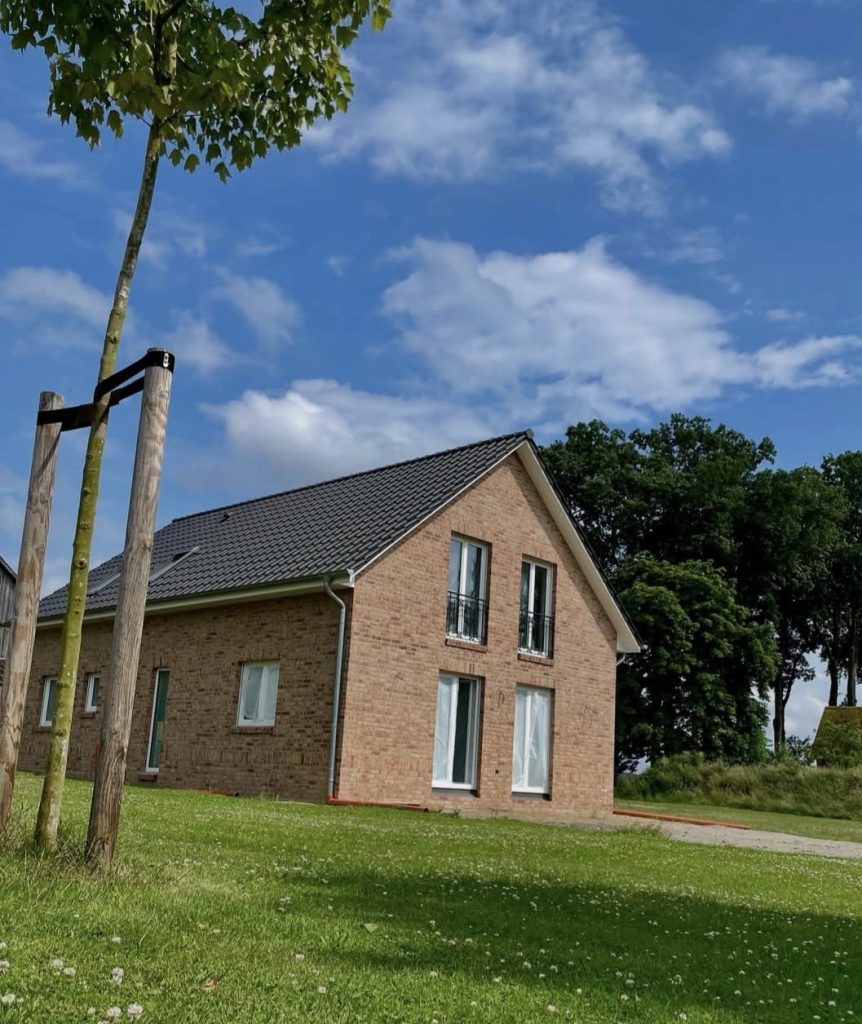 Fertiges, modernes Backsteinhaus mit dunkelgrauem Dach, weißen Fenstern und kleinen französischen Balkonen, umgeben von einer grünen Wiese und Bäumen.