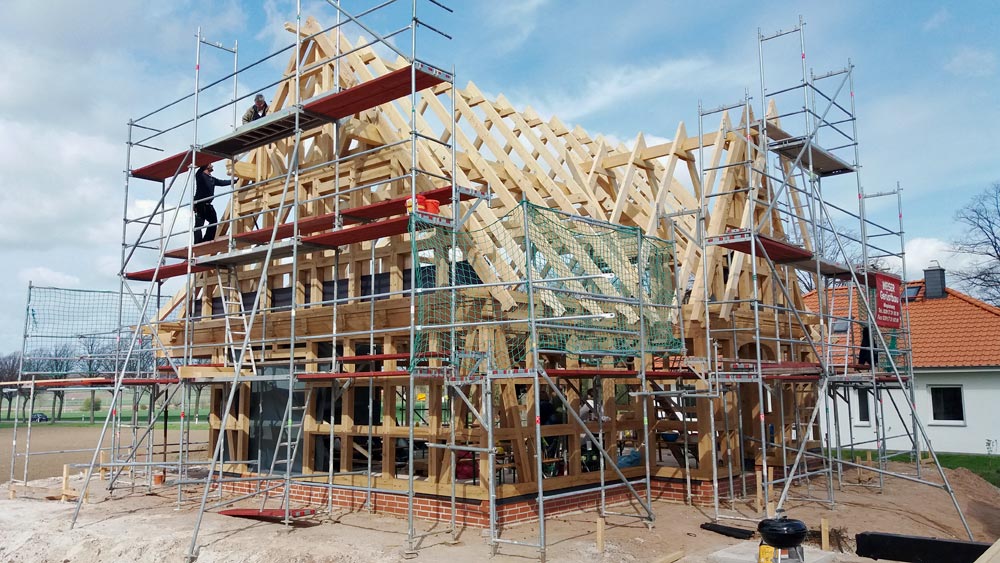 Fachwerkhaus im Bau, Holzrahmenstruktur und Dachstuhl sichtbar, umgeben von einem Baugerüst mit zwei Arbeitern.