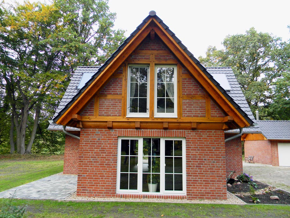 Vorderansicht eines fertiggestellten Hauses mit Backsteinmauerwerk im Erdgeschoss und Fachwerk im Giebel, mit weißen Sprossenfenstern und einem dunklen Ziegeldach.